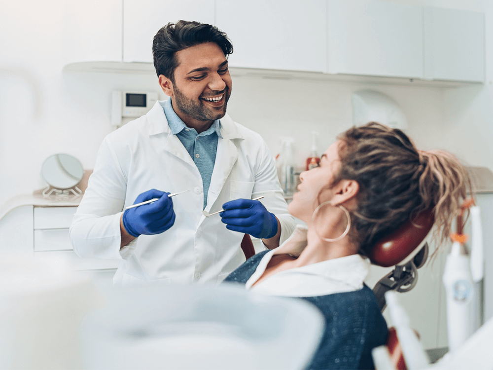 A male dentist wearing a white coat, blue gloves, and a blue shirt is smiling while holding dental tools and talking with a female patient. The patient, wearing a dark sweater and white collared shirt, is reclining in a dental chair, looking at the dentist. The background features a clean, modern dental office with equipment and cabinets.