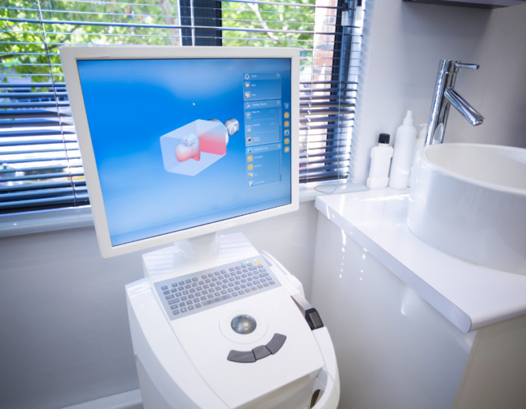 A dental CAD/CAM machine in a modern dental office, displaying a 3D digital model of a tooth restoration on its screen. The machine has a built-in keyboard and control panel. The background features a sink, bottles of dental supplies, and a window with blinds partially open, letting in natural light