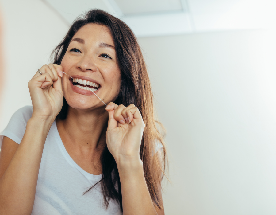 A woman with long dark hair and a white t-shirt is smiling while flossing her teeth. She is looking at her reflection in a mirror, holding the floss taut between her fingers. The background is softly lit and neutral-colored.