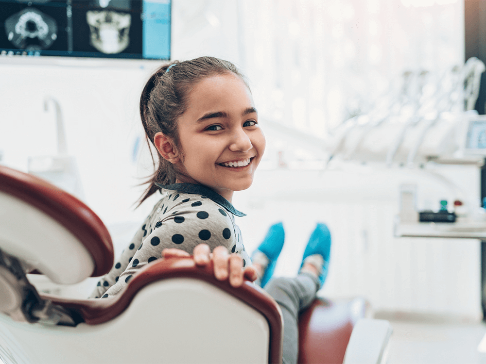 A young girl with dark hair tied in a ponytail is sitting in a dental chair, smiling and looking back at the camera. She is wearing a polka-dotted shirt, and her legs are stretched out with blue shoe covers on. The background shows a bright, modern dental office with dental equipment and an X-ray displayed on a screen.