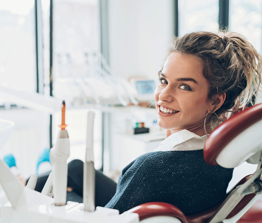 A young woman with wavy hair tied back is sitting in a dental chair, smiling and looking over her shoulder. She is wearing a dark sweater over a white collared shirt. The background features a bright and modern dental office with dental tools and equipment.