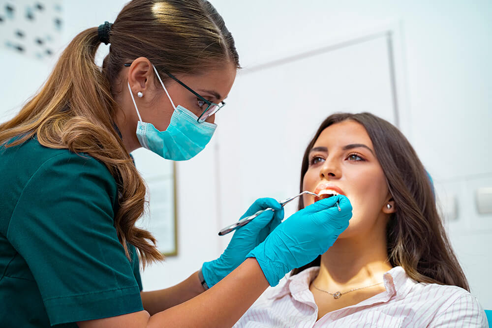 A female dentist or dental hygienist wearing a green uniform, protective mask, and gloves is examining a young woman's teeth using a dental mirror and scaler. The patient, wearing a white and pink striped shirt, sits in a dental chair with her mouth open while looking up.
