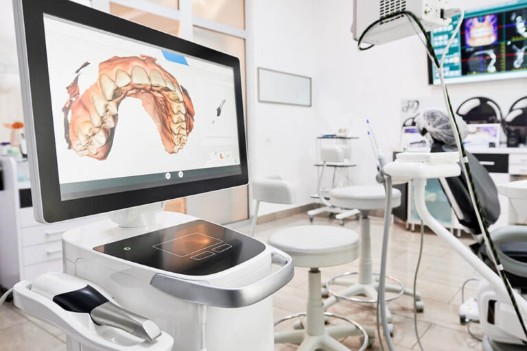 A close-up of an intraoral scanner in a modern dental office. The screen displays a 3D digital scan of a patient's upper teeth, while the scanner handpiece rests on the device. The background features a dental chair, stools, and other dental equipment.